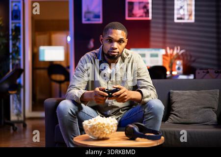 Der afroamerikanische Guy verbringt seine Freizeit mit einer Schüssel Popcorn und spielt während des Spielabends in seiner gemütlichen Wohnung Videospiele. Junger Erwachsener, der seine Konsole mit dem Fernseher verbindet. Stockfoto