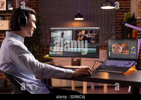 Fröhlicher Freelancer, der den Soundtrack zu Hause bearbeitet und Videos auf mehreren Geräten auf dem Schreibtisch vergleicht. Asiatischer Filmemacher, der visuelle Effekte, Ton- und Farbabstufungen während der Post-Production-Session überprüft. Stockfoto