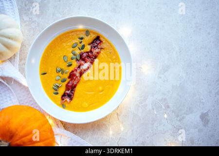 Cremige Kürbissuppe garniert mit knusprigem Speck und Kürbiskernen, serviert in einer weißen Schüssel auf strukturierter Oberfläche, umgeben von d Stockfoto