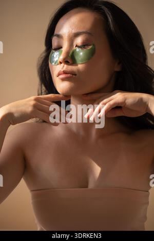 Asiatische Frau, die sich mit grünen Augenflecken im Gesicht entspannt und eine Hautpflege-Routine präsentiert, mit sanfter Beleuchtung und einer ruhigen Atmosphäre Stockfoto