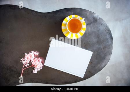 Flache Lay-Komposition mit weißem Notizbuch, einer Tasse Tee mit einer gestreiften gelben Untertasse und zarten rosa Blüten auf einem Moder Stockfoto