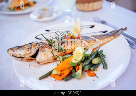 Gegrillter Fisch, serviert auf einem weißen Teller mit farbenfrohem Gemüse, garniert mit Kräutern, präsentiert ein köstliches Meeresfrüchte-Gericht in einem br Stockfoto
