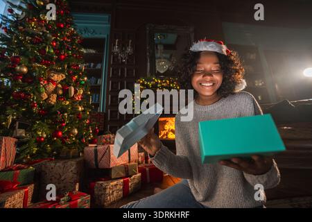 Frohe Weihnachten. Afroamerikanerin, die eine Geschenkbox in der Nähe des Weihnachtsbaums auspackt. Mädchen im Wohnzimmer mit Weihnachtsbaum Stockfoto