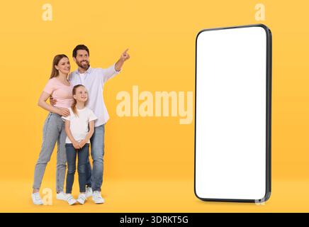 Familie, die ein großes Smartphone mit leerem Bildschirm auf orangem Hintergrund sieht. Vater zeigt auf Gerät Stockfoto