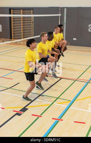 Verschiedene männliche Mannschaftskameraden führen synchronisierte Kniehegelungen in der Sporthalle mit Volleyballnetzpfosten durch. Athleten, Teamwork, Fitness, Stockfoto
