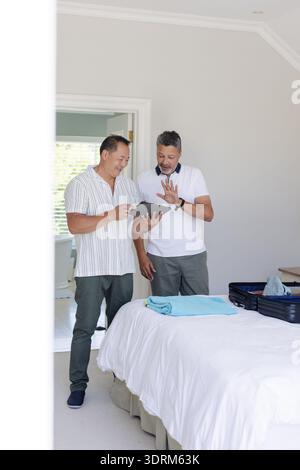 Zwei Männer im Schlafzimmer packen Koffer und diskutieren Reisepläne mit Tablet. Planung, Technologie, Vorbereitung, Urlaub, Adven Stockfoto