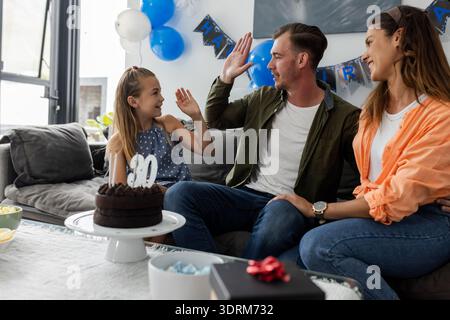 Wir feiern Geburtstag, die Familie gibt High Fives auf der Couch mit Kuchen und Ballons. Feier, Glück, Zweisamkeit, Party, che Stockfoto