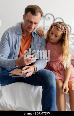 Vater und Tochter sitzen auf dem Bett, schauen zusammen auf das Smartphone und lächeln. Familie, Bindung, Technologie, Erziehung, Verbindung, Stockfoto