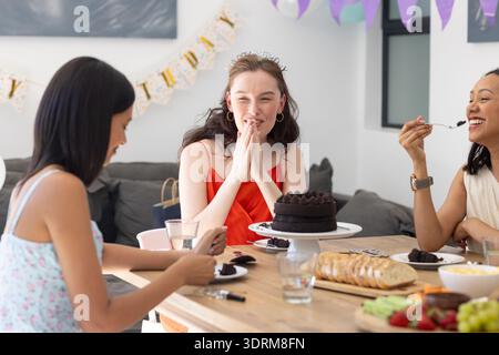 Lachen und genießen Kuchen, drei junge Frauen versammeln sich um einen Holztisch. Wenn sie Freundschaft feiern, teilen sie freudige Momente und Stockfoto