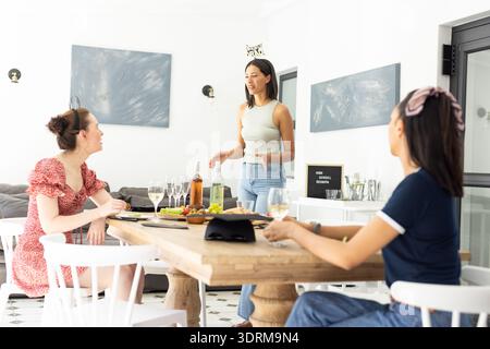 Drei junge Frauen treffen sich zu Hause um einen Holztisch. Sie feiern Freundschaft mit Getränken und Snacks Stockfoto