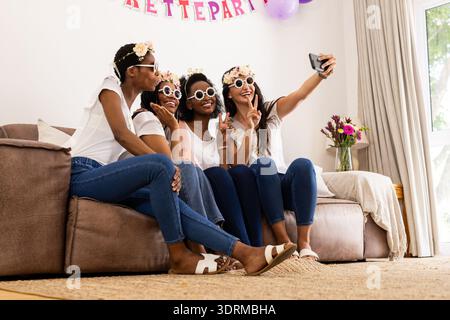 Auf der Couch sitzen, Freunde mit Blumenkronen, Selfie machen und zusammen lachen. Freundschaft, Freizeit, Bindung, Glück, fl Stockfoto