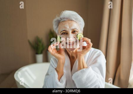 Reife Erwachsene alte Frau mit pflegender Anti-Age-Gesichtsmaske, die Gurkenscheiben hält. Ältere Frau mit weißer Maskencreme auf s Stockfoto