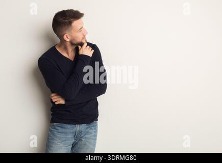 Der Mann sieht nachdenklich aus, während er neben einer Ebenen Wand in einer ungezwungenen Umgebung mit einer entspannten Haltung steht Stockfoto