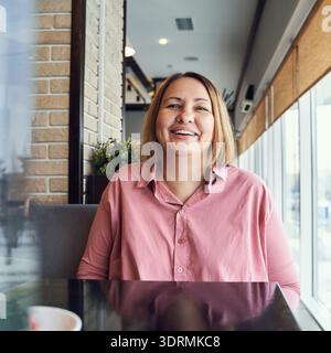 Kaukasische reife Frau lächelt im Café mit rosa Hemd drinnen Stockfoto