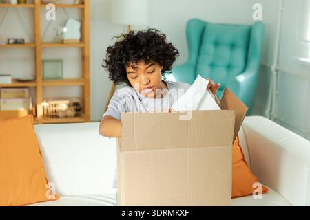 Afrikanisches Mädchen, das die Lieferung auspackt und in der Kiste sucht. Glückliche Frau, die den Karton öffnet. Eine Frau, die Pakete bekommt und die gelieferte Ware ansieht Stockfoto