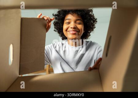 Innenseite des Kartons, Ansicht von unten. Afrikanisches Mädchen, das die Lieferung auspackt und in der Kiste sucht. Frau, die den Karton öffnet. Weibliche Paketbestellung Stockfoto