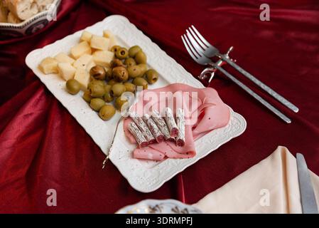 Weiße, geprägte rechteckige Servierplatte auf einer roten Tischdecke aus Stoff mit Würfeln aus hellem Käse, grünen Oliven, gefalteten Schinkenscheiben und kleinen Salami sti Stockfoto