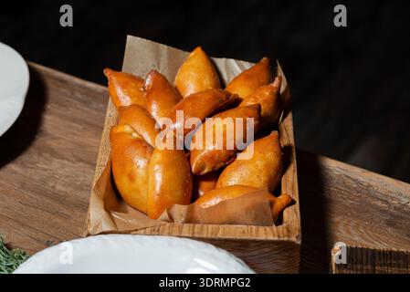 Ein rustikales Holztablett mit braunem Pergamentpapier ist gefüllt mit glänzenden, goldbraunen, frittierten Empanadas. Das Tablett sitzt auf einem Woo Stockfoto