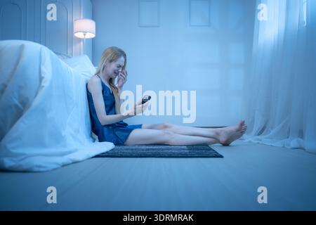 Eine junge Frau im blauen Pyjama fühlt sich traurig, nachdem sie Informationen von ihrem Handy erhalten hat Stockfoto