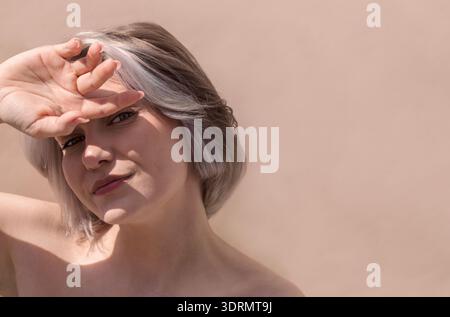 Junge Frau mit kurzen silbernen Haaren, die ihre Augen spielerisch vor Sonnenlicht schützt, zeigt natürliche Schönheit und weiche Hauttöne aga Stockfoto