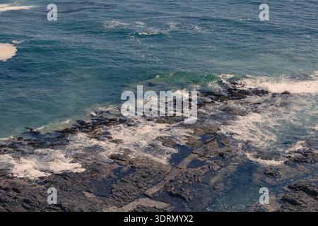Hochwinkelaufnahme türkisfarbener Pazifik-Wellen, die in Iquique, Chile, gegen dunkle vulkanische Felsen prallen, mit natürlichen Texturen und Morgenlicht Stockfoto