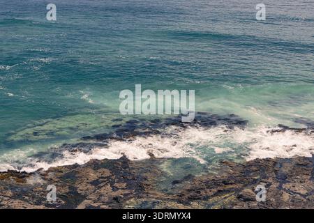 Hochwinkelaufnahme türkisfarbener Pazifik-Wellen, die in Iquique, Chile, gegen dunkle vulkanische Felsen prallen, mit natürlichen Texturen und Morgenlicht Stockfoto