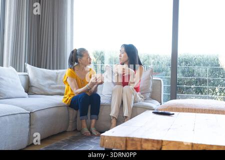 Asiatisch-chinesische Mutter und Tochter plaudern in hellem Wohnzimmer auf Sofa und halten weiße Tassen. Nähe, Wärme, Familie, Kompani Stockfoto
