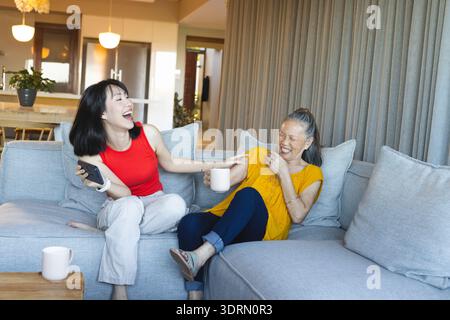 Asiatische Mutter und Tochter teilen sich Kaffeetassen und Smartphone im Wohnzimmer und lachen zusammen. Familie, Bindung, Freizeit, Kontem Stockfoto