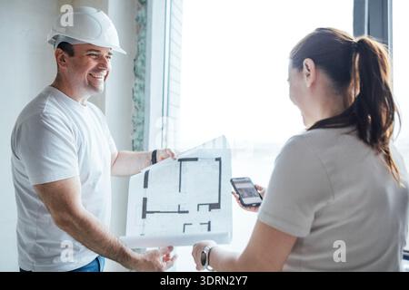 Der Bauarbeiter mit Schutzhelm lächelt, während er mit einem Kollegen Pläne bespricht und Teamarbeit und Zusammenarbeit in einem vorstellt Stockfoto