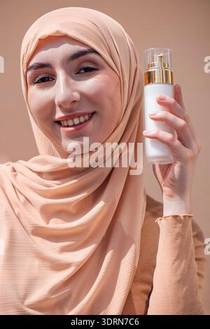 Eine junge Frau, die einen beigen Hijab trägt, hält eine Hautpflegeflasche, lächelt selbstbewusst und zeigt Schönheitsprodukte in einer weichen Neutra Stockfoto