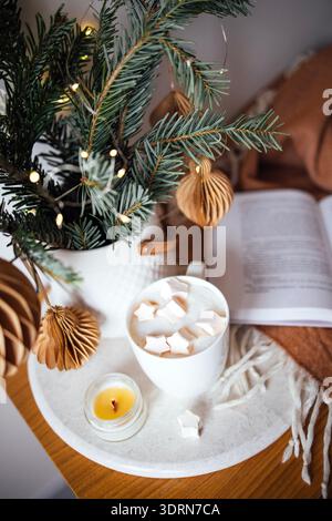 Gemütliche Winterszene mit einem weißen Becher voller Marshmallows, umgeben von festlichen Dekorationen, einer Kerze und einem Buch, Kreation Stockfoto