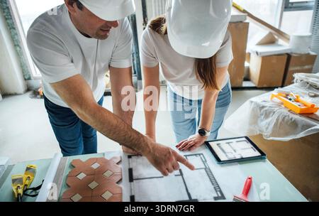 Zwei Bauprofis, ein Mann und eine Frau, die Schutzhelme tragen, diskutieren Architekturpläne auf einem Tisch in einem modernen Stockfoto