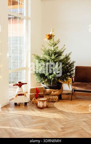 Der mit einem Stern dekorierte Weihnachtsbaum steht in einem gemütlichen Wohnzimmer, umgeben von festlicher Dekoration, einem bequemen Stuhl und W Stockfoto