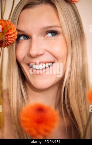 Lächelnde Frau mit langen blonden Haaren, umgeben von leuchtenden orangefarbenen Blumen, die einen fröhlichen Ausdruck und natürliche Schönheit in A zeigt Stockfoto