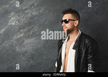 Stylische Mann in schwarzer Lederjacke posiert vor grauem Hintergrund mit Sonnenbrille Stockfoto