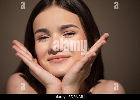 Eine junge Frau mit langen dunklen Haaren, die lächelnd ihr Kinn auf den Händen hält und einen fröhlichen Ausdruck in einem weichen, warmen Licht zeigt Stockfoto