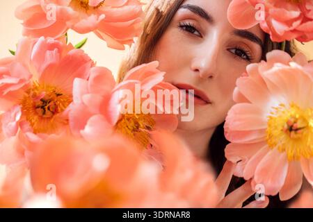 Junge Frau mit langen braunen Haaren umgeben von leuchtenden rosa Pfingstrosen, die natürliche Schönheit und florale Eleganz in einem weichen, dr. Stockfoto
