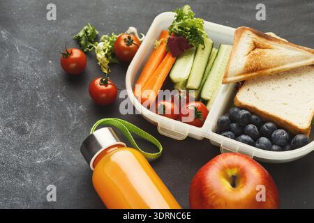 Mittagessen mit frischem Gemüse, Obst und Getränk in einer Lunchbox auf dunklem Hintergrund Stockfoto