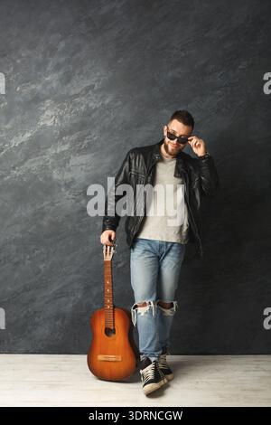 Der Mann steht mit einer Gitarre vor einer dunklen Wand, trägt eine Sonnenbrille und eine Lederjacke Stockfoto