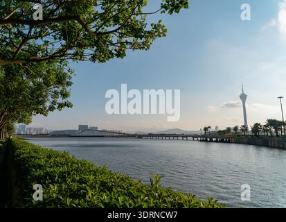 Der Macau Tower steht hoch über einer langen Brücke, die ruhiges Wasser unter klarem blauem Himmel mit üppigem Grün umrahmt. Stockfoto