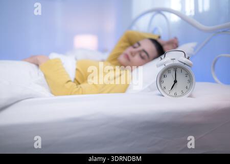 Ein weißer Wecker auf einem Bett mit einer Frau, die im Hintergrund schläft, veranschaulicht die morgendlichen Routinen und die friedliche Ruhe Stockfoto