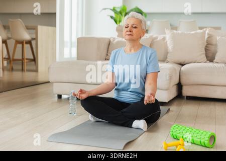 Yoga-Achtsamkeitsmeditation. Ältere Erwachsene reife Frau, die zu Hause Yoga praktiziert. Die alte Dame im mittleren Alter sitzt in Lotusposition auf Yoga M Stockfoto