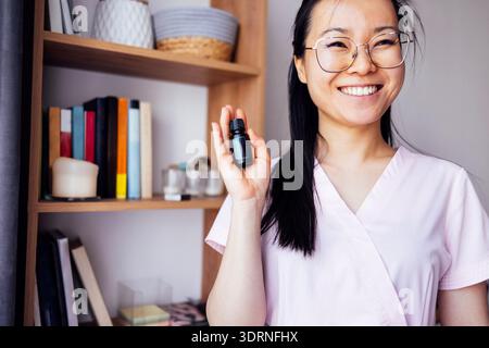 Asiatische Frau in rosafarbenen Schutt hält eine kleine Flasche ätherisches Öl und lächelt hell in einem gemütlichen Raum mit Regalen voller Bücher Stockfoto