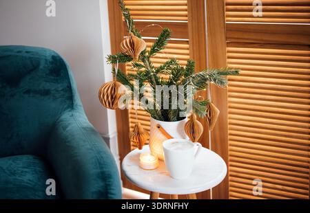 Gemütliche Wohnzimmerszene mit einem dekorativen immergrünen Arrangement mit Papierornamenten, einer warmen Kerze und einem weißen Becher auf einer Mutter Stockfoto