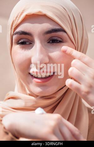 Eine junge Frau, die einen beigen Hijab trägt, trägt Hautpflegecreme auf ihr Gesicht auf und zeigt eine Schönheitsroutine in einem hellen Outdoor-Set Stockfoto