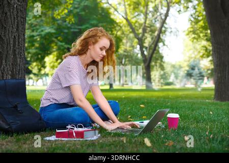 Der Schüler arbeitet am Laptop im Park während des hellen Sonnentages mit Kaffee und Rucksack in der Nähe Stockfoto
