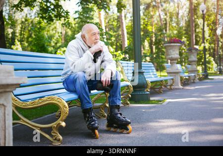 Ein älterer Mann ruht sich tagsüber auf einer Bank aus, während er Rollschuhe in einem Park trägt Stockfoto