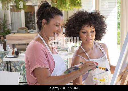 Asiatische Mutter und afroamerikanische Tochter malen an der Staffelei im Hinterhof auf bespritzten Schürzen. Kreativ, Freizeit, unterwegs Stockfoto