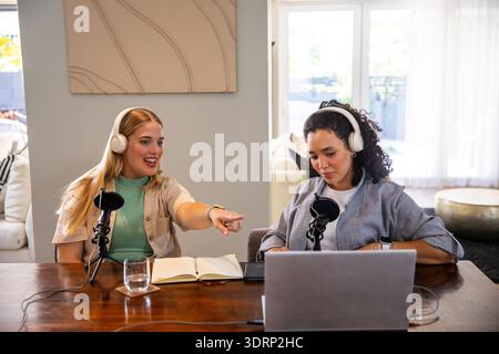 Verschiedene weibliche Co-Moderatoren, die Podcasts im Wohnzimmer an einem Holztisch mit Laptop-Mikrofonen aufzeichnen. Podcast, Aufnahme, Co-Hosts, Stockfoto
