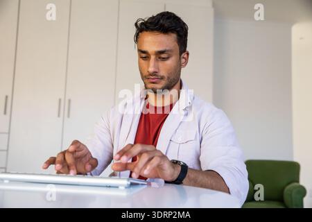 Asiatischer Mann, der am weißen Schreibtisch sitzt und im Heimbüro in der Nähe von Aufbewahrungsschränken auf der kabellosen Tastatur schreibt. Konzentration, Produktivität, m Stockfoto